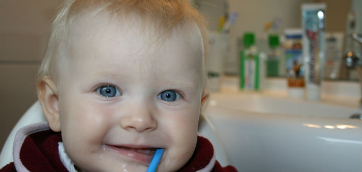 A blond baby with big blue eyes dangles a blue toothbrush from his mouth.