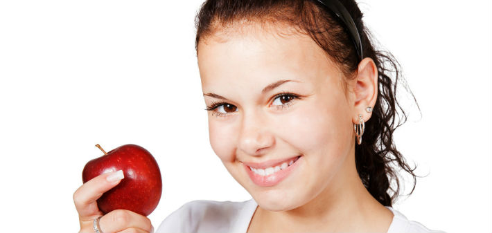 A dark haired woman smiles and holds a red apple in front of a white background.