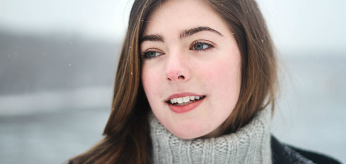 A pale, dark haired woman dressed for winter stands in the snow, smiling gently against a pale, blurred background.