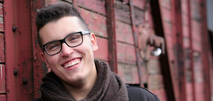 A teen boy with glasses smiles widely, showing well cared for teeth as he leans against a red, wooden wall.