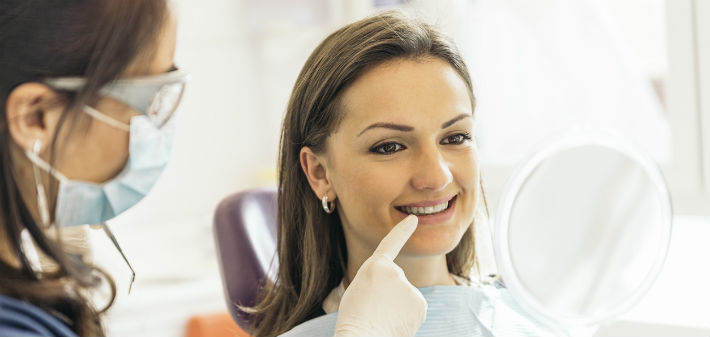 A female patient smiles into a handheld mirror as a masked female dental technician points to the patient's tooth with one finger.