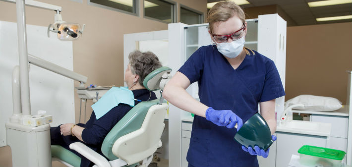 A female technician mixes material to make a mold for dentures behind an older, female patient siting in the dental chair.