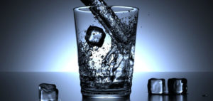 Water splashes into a clear glass with ice cubes, and ice cubes lay to either side against a blue background.
