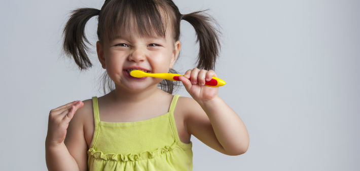 A little girl with pigtails looks thrilled as she brushes with a yellow toothbrush.