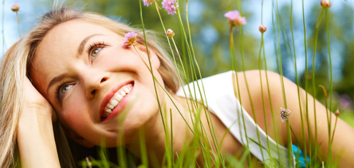 A young blonde woman lies in the grass on her stomach, head in hand, as she gazes upward and smiles with percelain veneers.