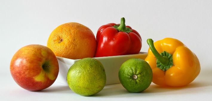 An orange and a red pepper sit in a white dish behind an apple, two lines, and a yellow pepper.