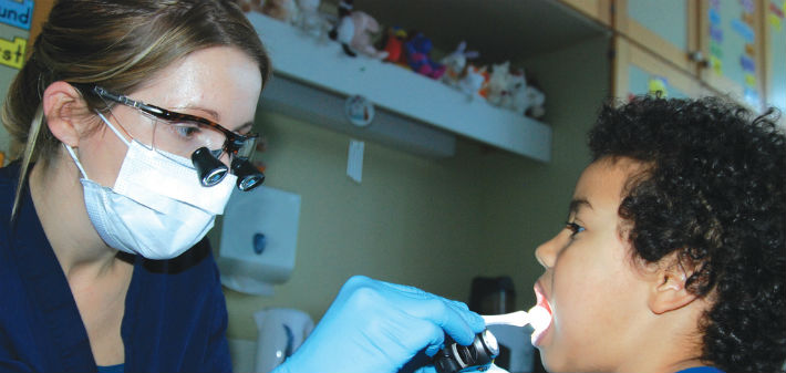 A female technician wearing binocular loupes and a mask uses a dental mirror and light to look in a young boy's mouth.