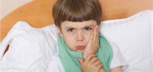 A boy in bed holds his cheek to protect a tooth that is sore from one of the common dental problems in children.