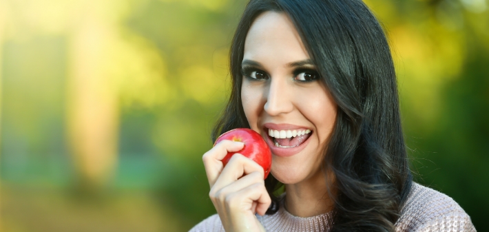 A woman holds a red apple, smiling and showing her healthy teeth.