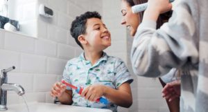 A mother and her son smile at each other while getting ready to brush their teeth with a second, mostly off-camera son.