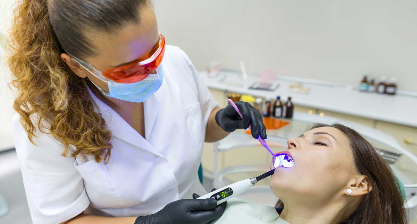 A female dentist looks into a comfortably sedated female patient's mouth with a dental mirror while curing a filling.