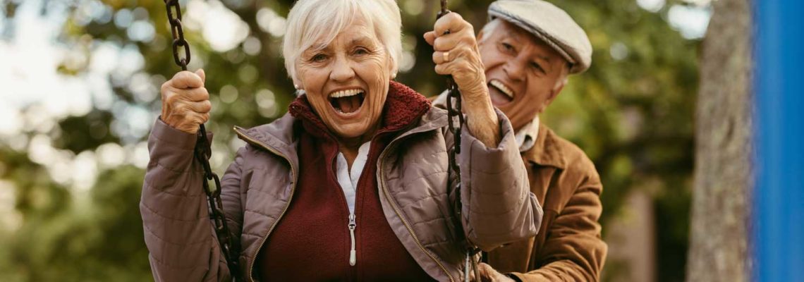 An older couple play on a swing, smile broadly to show off their mini dental implants.