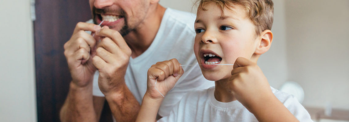 A father flosses his teeth with his son to show the boy the importance of flossing.