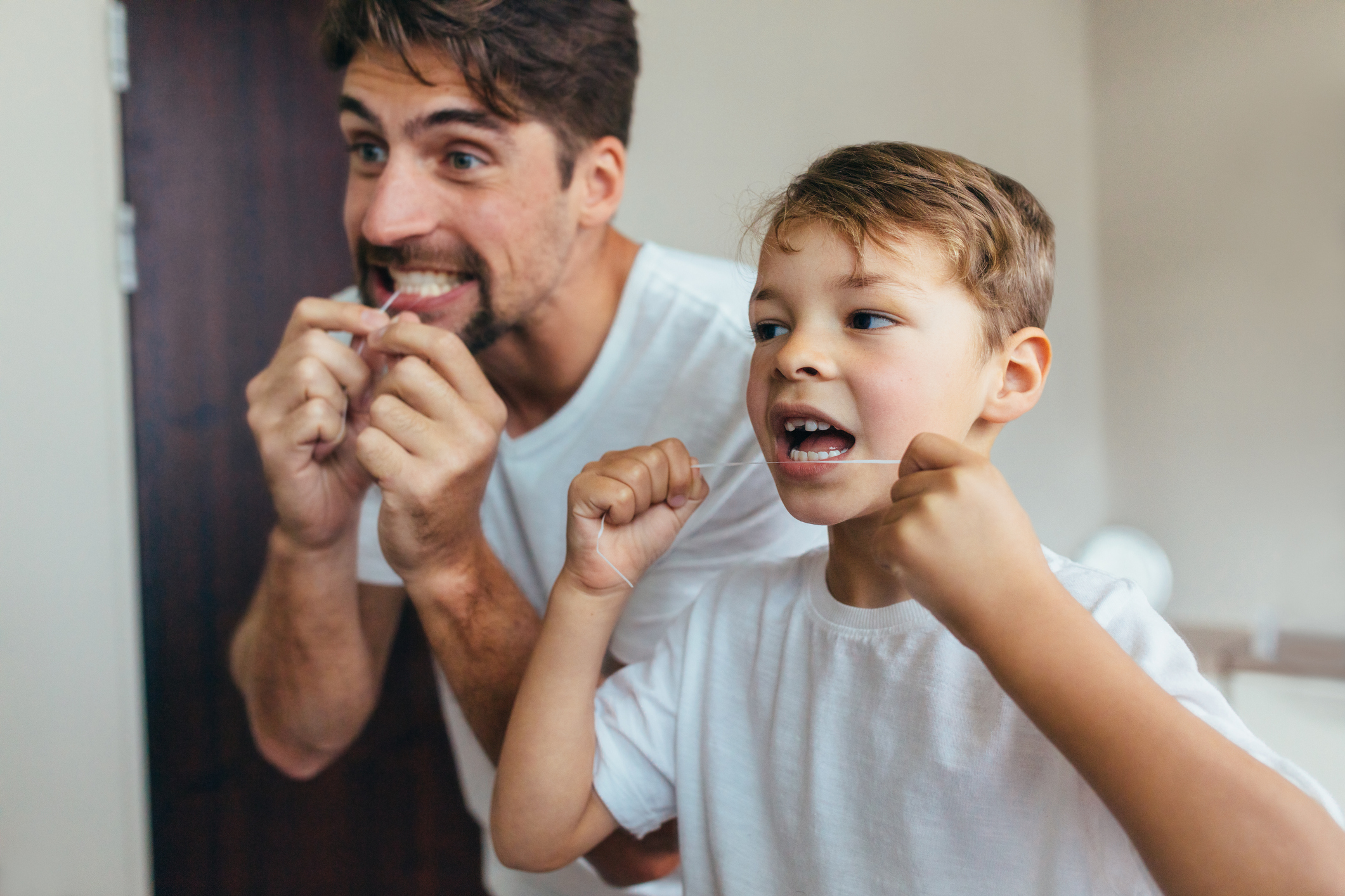 A father flosses his teeth with his son to show the boy the importance of flossing.