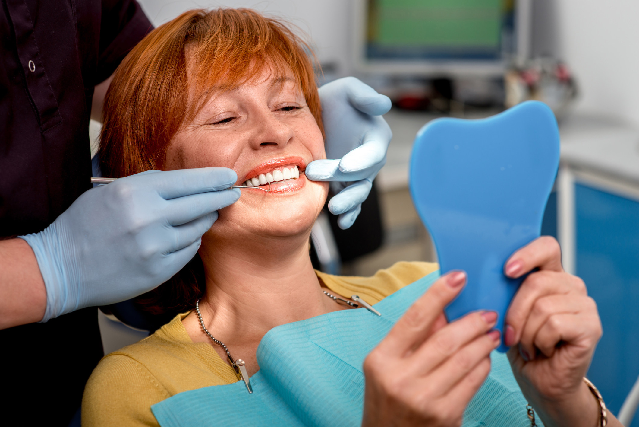 An older female patient smiles into a mirror as a technician explains the advantages of dental implants.
