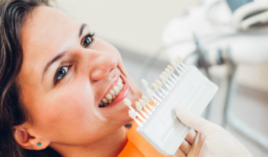 Model teeth in varying shades are held to a female patient's smile to choose the closest color for a cosmetic procedure.