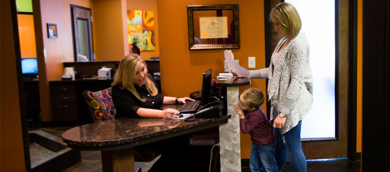 A female family-dentist receptionist smiles at a young boy standing beside his mother, who is reading a list of payment options.